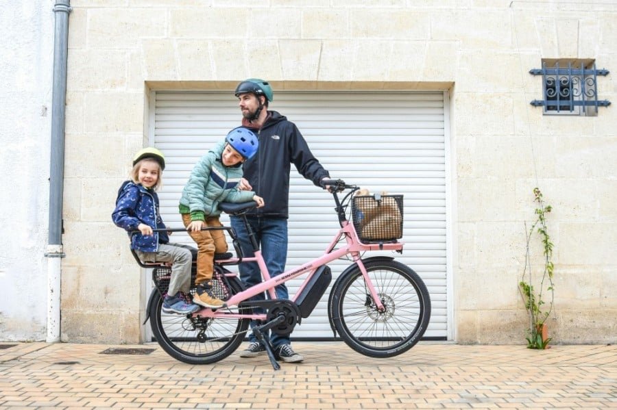 un homme et deux enfants sur un vélo cargo rose